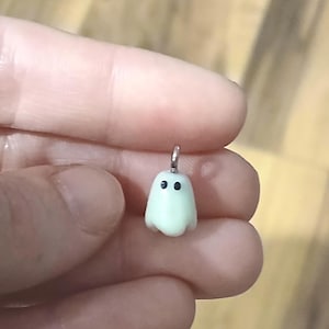 May include: A small, pale, off-white ghost-shaped charm with two simple black dots for eyes. The charm features a silver-toned metal jump ring attached to the top, allowing it to be suspended or attached to a chain or other jewelry component.