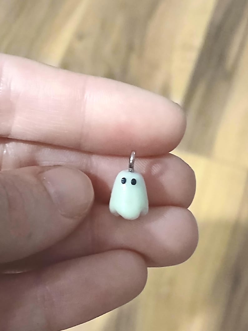 May include: A small, pale, off-white ghost-shaped charm with two simple black dots for eyes. The charm features a silver-toned metal jump ring attached to the top, allowing it to be suspended or attached to a chain or other jewelry component.