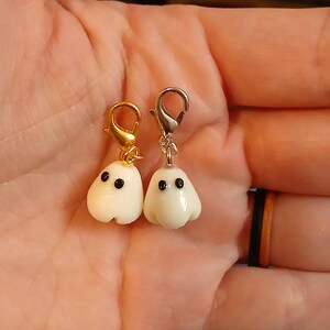 May include: Two small ghost charms made of smooth white material with black dot eyes. One ghost is attached to a gold-colored metal lobster clasp, and the other ghost is attached to a silver-colored metal lobster clasp for easy attachment.