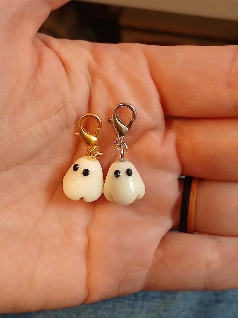 May include: Two small ghost charms made of smooth white material with black dot eyes. One ghost is attached to a gold-colored metal lobster clasp, and the other ghost is attached to a silver-colored metal lobster clasp for easy attachment.