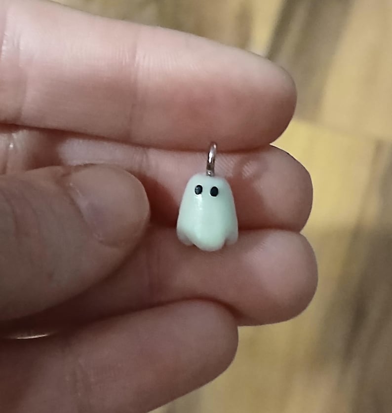 May include: A small, pale, off-white ghost-shaped charm with two tiny black dots for eyes. The charm features a smooth, rounded surface and includes a small silver-toned metal jump ring attached to the top for hanging on a necklace or bracelet.