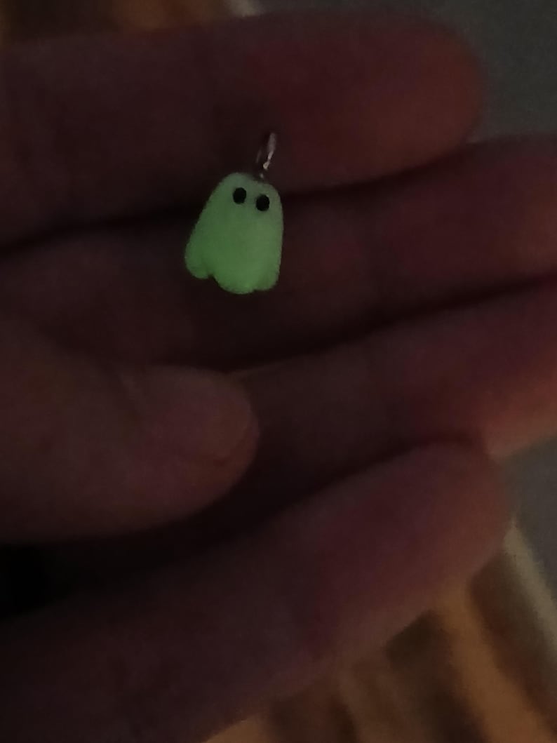 May include: A small, pale green ghost-shaped pendant featuring two simple black circular eyes. The charm includes a small silver-toned metal jump ring at the top, designed for attachment to a necklace chain or a bracelet, showcasing a smooth finish.