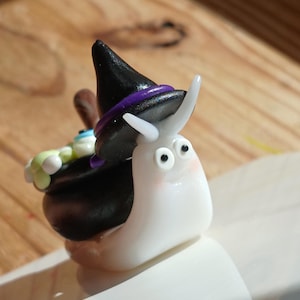 May include: A small white clay snail figurine wearing a black witch hat with a purple band. The snail carries a black cauldron on its back filled with small green and white spheres and a single blue eye, featuring two black eyes.