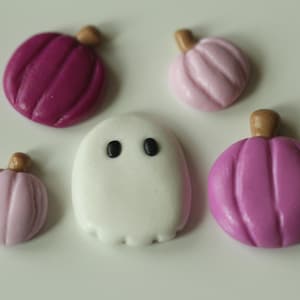 May include: Five Halloween-themed fondant decorations: a white ghost with black eyes, a pink pumpkin with a brown stem, a purple pumpkin with a brown stem, and two smaller pink pumpkins with brown stems.