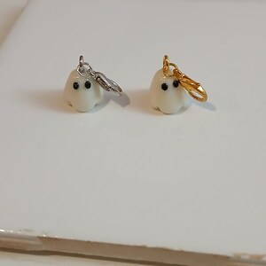 May include: Two small, off-white ghost charms with smooth surfaces and two black dot eyes. One ghost features a silver-toned metal lobster clasp attached to a jump ring, while the other ghost features a gold-toned metal lobster clasp attached to a jump ring.