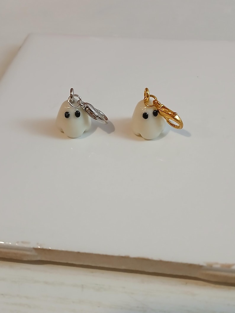 May include: Two small, off-white ghost charms with smooth surfaces and two black dot eyes. One ghost features a silver-toned metal lobster clasp attached to a jump ring, while the other ghost features a gold-toned metal lobster clasp attached to a jump ring.