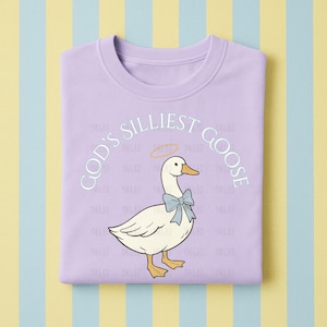 Może przedstawiać: Liliowa bluza z kapturem z kresk&oacute;wkową gęsią z niebieską kokardą i aureolą. Tekst "GOD'S SILLIEST GOOSE" jest wygięty nad gęsią. Bluza jest złożona na paskowanym tle.