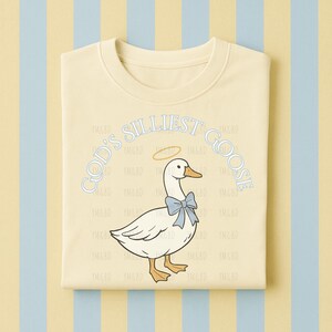 Może przedstawiać: Jasnoż&oacute;łta bluza z napisem "GOD'S SILLIEST GOOSE" w jasnoniebieskim kolorze. Grafika przedstawia białą gęś z niebieską muszką i aureolą. Bluza jest złożona na paskowanym tle.