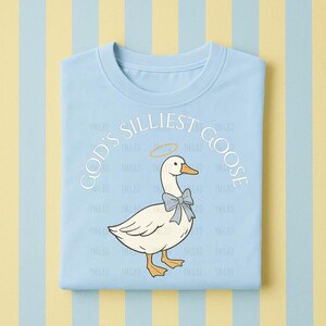 Może przedstawiać: Jasnoniebieska koszulka z napisem "GOD'S SILLIEST GOOSE" w okrągłym wzorze. Grafika przedstawia białą gęś z niebieską muszką i aureolą. Koszulka jest złożona na paskowanym tle.