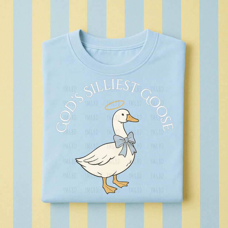 Może przedstawiać: Jasnoniebieska koszulka z napisem "GOD'S SILLIEST GOOSE" w okrągłym wzorze. Grafika przedstawia białą gęś z niebieską muszką i aureolą. Koszulka jest złożona na paskowanym tle.