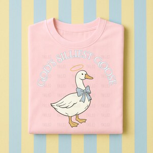 Może przedstawiać: Jasnor&oacute;żowa bluza z okrągłym dekoltem z napisem "GOD'S SILLIEST GOOSE" nad kresk&oacute;wkową gęsią w niebieskiej muszce i aureoli. Gęś jest biała z ż&oacute;łtymi stopami i dziobem. Tło jest w paski.