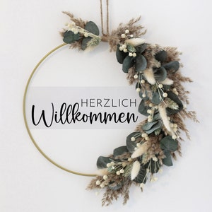 Peut inclure: Une couronne en métal doré décorée de verdure séchée, y compris de l'eucalyptus et de l'herbe à pampas. La couronne porte le texte "HERZLICH Willkommen" en noir.