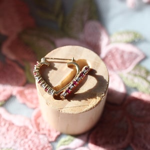 May include: A gold-coloured heart-shaped earring with red and clear gemstones. The earring is displayed on a small, light-coloured wooden cylinder, set against a backdrop of pink floral fabric.