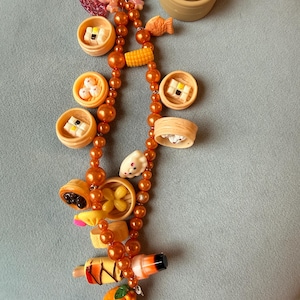 May include: A food-themed keyring with miniature dim sum and other Asian cuisine items. The charm includes a dumpling, fish, corn, and orange beads, all in shades of orange and brown. The keyring is attached to a silver clasp.
