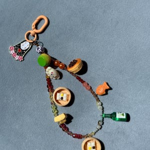 以下が含まれることがあります： ピーチカラーのロブスタークラスプと、ピンクの花をあしらった白い顔のキャラクターのメタルチャームが付いたキーホルダー。チェーンには、点心が入った竹製の蒸し器、緑色のボトル、魚など、さまざまなミニチュアフードチャームが飾られています。
