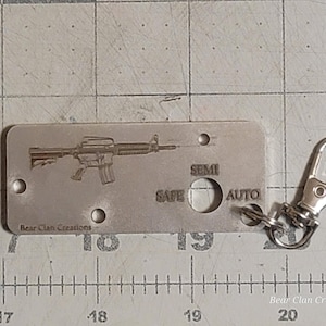 Könnte beinhalten: Ein silberner Schlüsselanhänger mit einem lasergravierten Bild eines Gewehrs und den Worten "SAFE", "SEMI" und "AUTO". Der Schlüsselanhänger wird von Bear Clan Creations hergestellt.