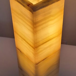 May include: A rectangular onyx lamp with a warm, glowing interior. The lamp's layered design features bands of yellow and cream hues, creating a natural, textured appearance. The top opening allows light to emanate, enhancing the lamp's aesthetic.