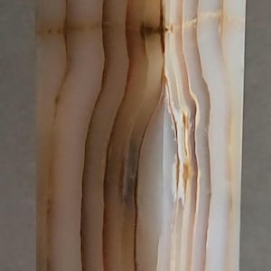 May include: A tall, rectangular pillar made of polished stone with a smooth, translucent surface. The object features vertical bands of cream, beige, and warm brown colours, creating a natural layered pattern throughout the entire structure.