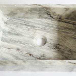 May include: A rectangular bathroom sink carved from natural white marble with prominent grey and charcoal veining throughout the stone. The basin features a smooth, polished interior surface and a centred circular drain hole for plumbing installation.