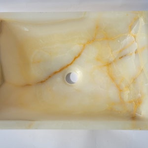 May include: Rectangular onyx sink in light yellow and cream tones. The sink has a central drain hole and natural gold and brown veining. The basin has a smooth, polished surface, ideal for a bathroom.