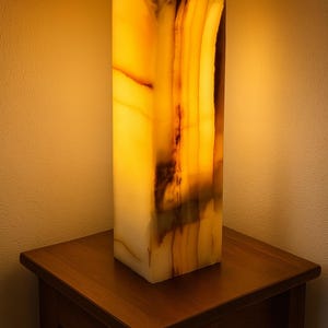 May include: A rectangular onyx lamp with a warm, glowing interior. The lamp is a light amber colour with darker brown veining. It sits on a small, dark wooden table. The lamp's dimensions are approximately 15 cm wide, 15 cm deep, and 30 cm tall.