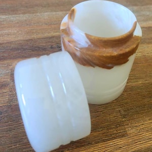 Puede incluir: Jarrón de ónix blanco con un patrón de remolino marrón. El jarrón tiene una tapa con un acabado liso y blanco.