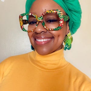 May include: A woman wearing a green head wrap, large patterned glasses, and flower-shaped earrings. She is wearing a yellow turtleneck. The glasses and earrings have a colorful, patterned design.