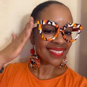 May include: A woman wearing a bright orange shirt with large, colorful, patterned earrings and glasses. The glasses are also patterned with a similar design to the earrings.