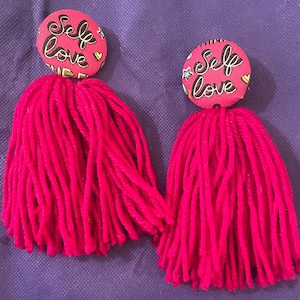 May include: A pair of pink tassel earrings with a pink button featuring the text "Self Love" in black lettering.