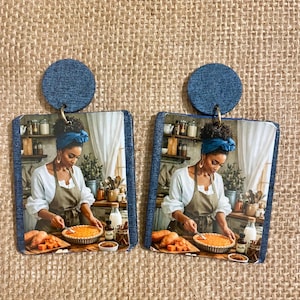May include: Earrings with a square image of a woman baking a pie, topped with a denim blue circle. The woman wears a white shirt, apron, and headscarf. The earrings are set against a burlap background, with a rustic aesthetic.