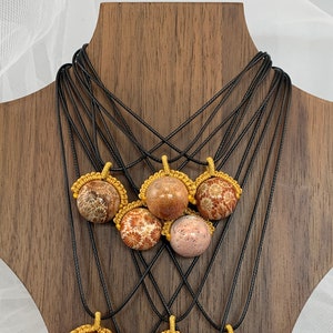 Puede incluir: Una muestra de ocho collares con colgantes de piedra de color coral. Cada colgante est&aacute; rodeado por un cord&oacute;n anudado de color dorado. Los collares se muestran en un soporte de madera.