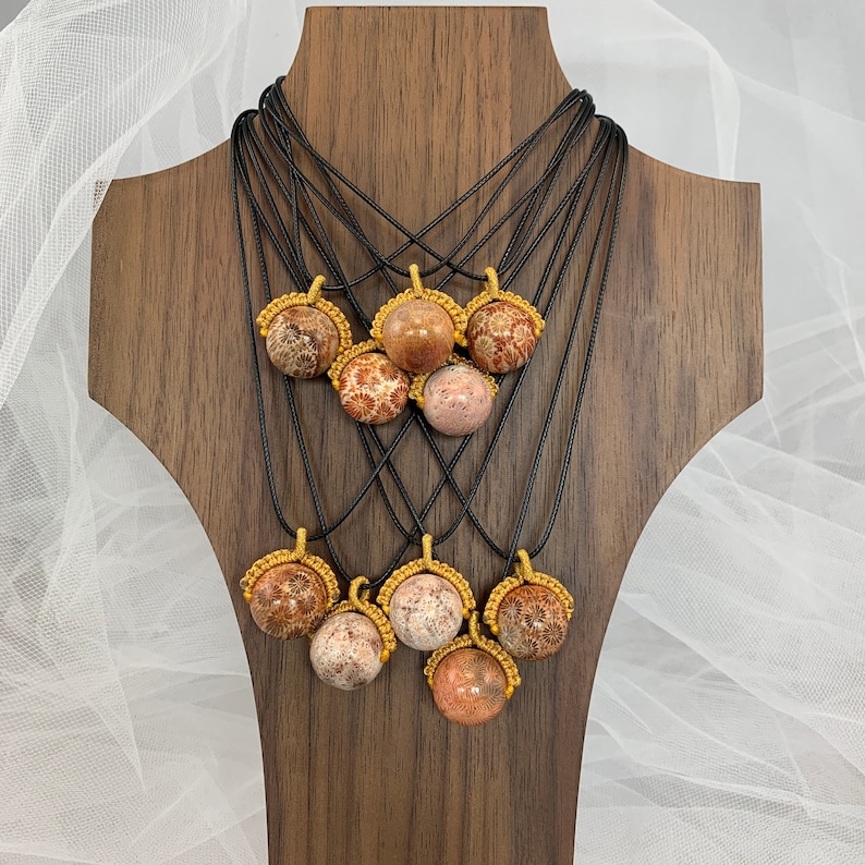 Puede incluir: Una muestra de ocho collares con colgantes de piedra de color coral. Cada colgante est&aacute; rodeado por un cord&oacute;n anudado de color dorado. Los collares se muestran en un soporte de madera.