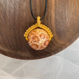 Puede incluir: Un collar con un colgante esf&eacute;rico de color dorado y marr&oacute;n con un patr&oacute;n floral y un cord&oacute;n negro.