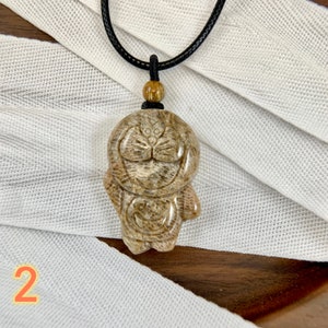 May include: A brown and white carved stone pendant of a cartoon character with a black cord necklace. The pendant is on a white fabric background.