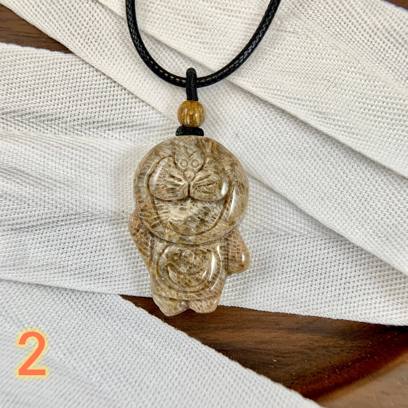 May include: A brown and white carved stone pendant of a cartoon character with a black cord necklace. The pendant is on a white fabric background.