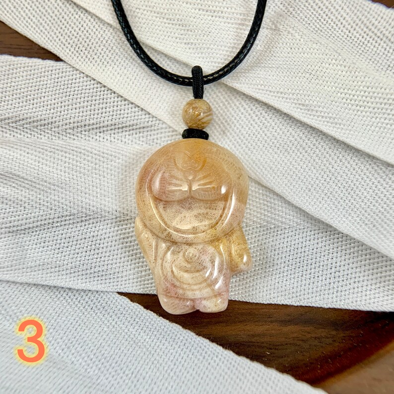 May include: A tan and white carved stone pendant of a cartoon character with a black cord necklace. The pendant is shaped like a person with a round head and a wide smile.