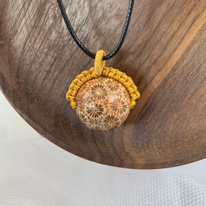 Puede incluir: Un collar con un colgante redondo de piedra con estampado floral dorado y marr&oacute;n. El colgante est&aacute; suspendido de un cord&oacute;n negro.