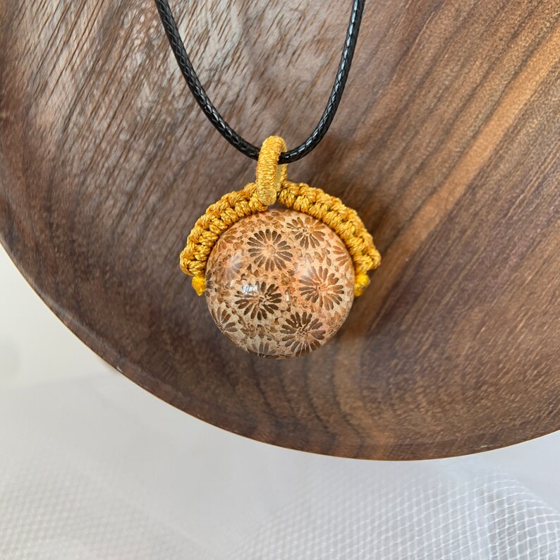 Puede incluir: Un collar con un colgante redondo de piedra con estampado floral dorado y marr&oacute;n. El colgante est&aacute; suspendido de un cord&oacute;n negro.