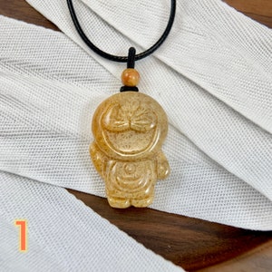 May include: A beige, carved stone pendant in the shape of a smiling cartoon character with a black cord necklace. The pendant has a detailed design with a textured surface.