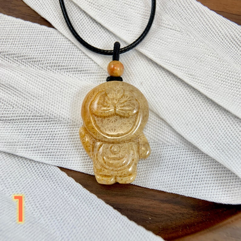 May include: A beige, carved stone pendant in the shape of a smiling cartoon character with a black cord necklace. The pendant has a detailed design with a textured surface.