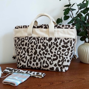 May include: A tote bag with a leopard print pattern in brown and beige. The bag has beige handles and trim. A ball of yarn, a knitted item, and a plant in a white vase are also on the wooden surface.