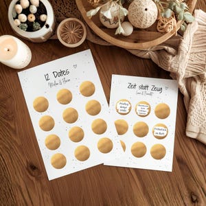 Könnte beinhalten: Zwei weiße Karten mit goldenen Kreisen, eine mit der Aufschrift "12 Dates" und die andere mit "Zeit statt Zeug". Die Karten liegen auf einer Holzoberfläche, mit dekorativen Gegenständen im Hintergrund. Die Karten haben Text auf Englisch und Deutsch.