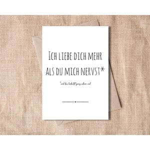 Könnte beinhalten: Eine weiße Grußkarte mit schwarzem Text, der lautet: "ICH LIEBE DICH MEHR ALS DU MICH NERVST*" und "*und das bedeutet ganz schon viel". Die Karte liegt auf einem braunen Umschlag, vor einem strukturierten, beigen Hintergrund.