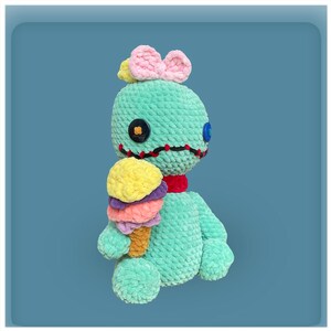 May include: A teal blue crocheted plush toy with a pink bow on its head, holding a multi-colored ice cream cone. The toy has black stitching around its eyes and mouth, and two button eyes.