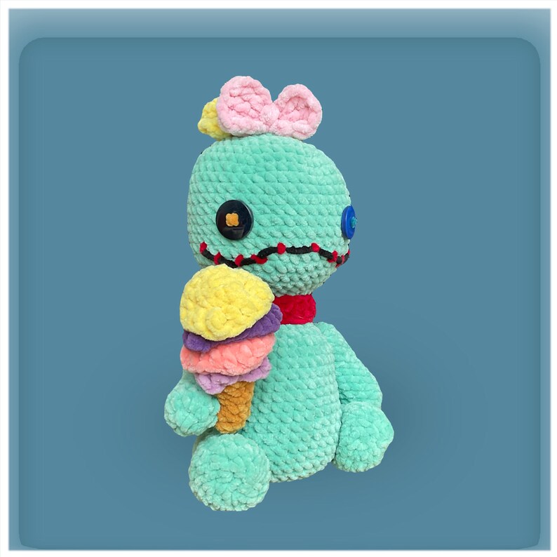 May include: A teal blue crocheted plush toy with a pink bow on its head, holding a multi-colored ice cream cone. The toy has black stitching around its eyes and mouth, and two button eyes.
