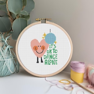 May include: A cross-stitch embroidery hoop with a white background and a pink heart with a smiling face holding a blue disco ball. The text "It's ok to dance alone" is stitched in green below the heart.