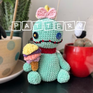 May include: A teal blue crocheted Stitch plush toy holding a multi-colored ice cream cone. Stitch is wearing a red scarf and a pink bow. The toy is sitting on a table with a cup and saucer and a potted plant in the background.