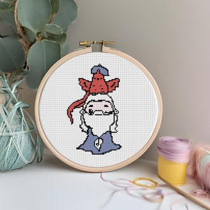 May include: A cross-stitch embroidery hoop with a white background and a cartoon image of a person with a red and blue bird on their head. The person is wearing a blue shirt and has a white beard.