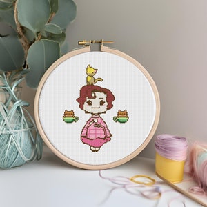 Può includere: Un cerchio da ricamo a punto croce con sfondo bianco e una donna vestita di rosa con i capelli castani e un gatto giallo sulla testa. Due gatti marroni con tazze verdi sono ai lati di lei.