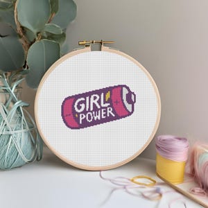 Puede incluir: Un bastidor de bordado de punto de cruz con un diseño de batería púrpura y las palabras "GIRL POWER" en blanco. La batería tiene acentos rosas y amarillos. El bastidor está sobre un fondo blanco.